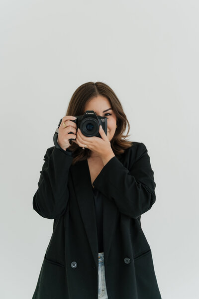 Smiling woman in a black blazer and jeans holding a camera up to her face in a minimal studio setting.