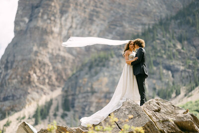This captivating wedding photograph captures the bride and groom holding each other at Moraine Lake in Banff National Park. With the bride's veil flowing high in the wind and the stunning lake and mountains behind them, the moment is filled with romance and natural beauty.