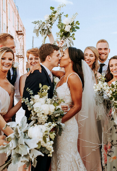Bride and groom kiss while wedding party surround them and cheer in alleyway