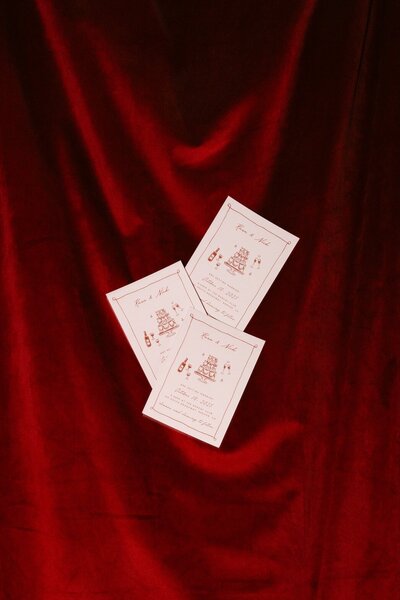 Creative wedding flatlay of illustrated invitations thrown into the air in front of a deep red velvet curtain at a Denver, Colorado dive bar wedding