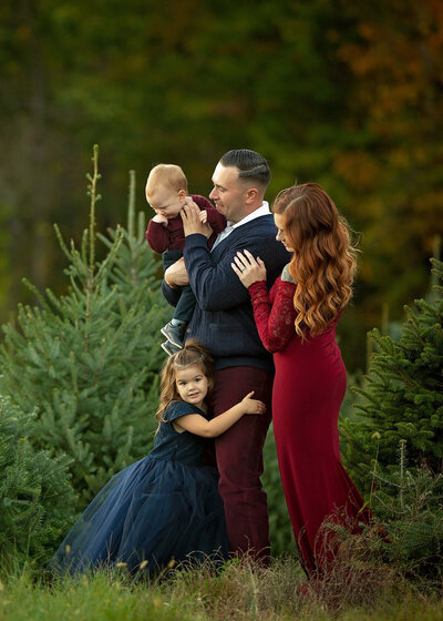 A husband and wife are standing with their son and daughter on a tree farm, wearing all blue and red