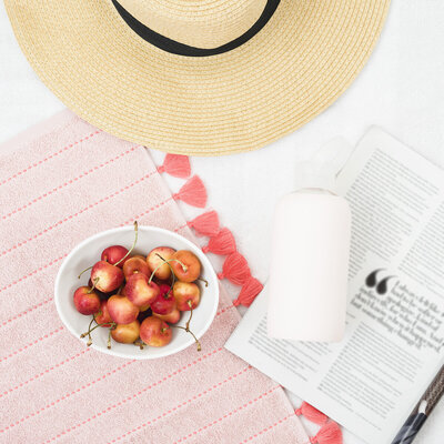 Cherries in a white bowl on top of a coral towel next to a sunhat and water bottle.