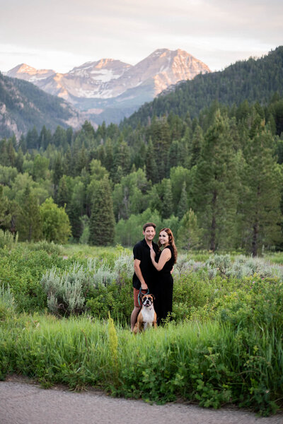 A man, woman, and their dog standing and smiling at the camera in front of a beautiful mountain backdrop