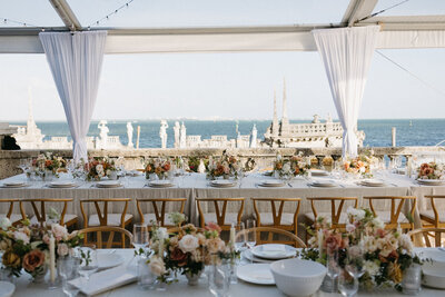 An outdoor wedding reception setup overlooking the water. The tablecloths are beige, the chairs are wooden with ivory cushions, and the centerpieces are a mix of peach, orange, and red florals