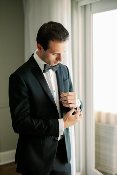 groom standing next to a window adjusting the sleeve of his wedding day tuxedo