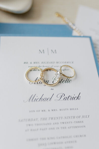 The wedding rings and engagement ring lay on top of a wedding invitation over the couple's names