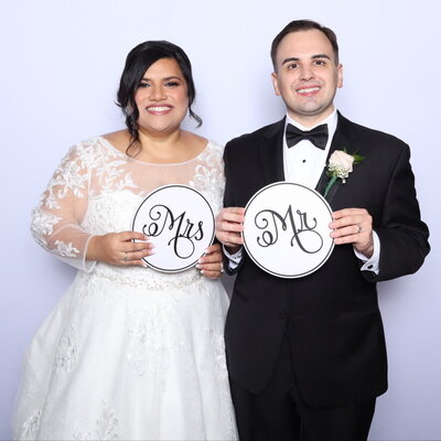 Bride and groom dressed in wedding attire holding circular “Mr” and “Mrs” signs in front of a light-colored photo booth backdrop.