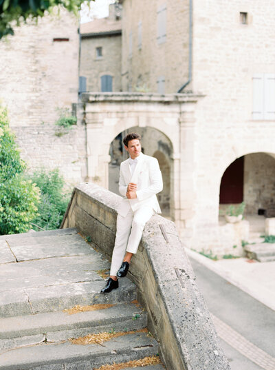 Groom sits on stone railing of staircase in old European town