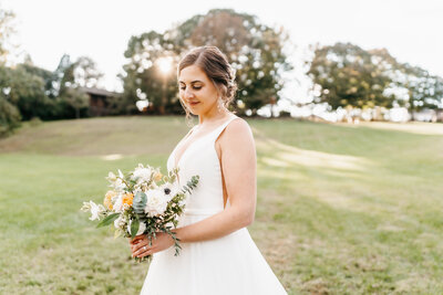 A bride in a white wedding dress holding a bouquet stands in a field during sunset