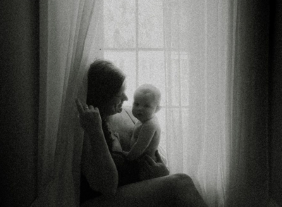 A mother and baby sit by a window. The mother is looking at the baby, the baby is looking at the camera while in their mother's arms. The mother is holding the window curtain