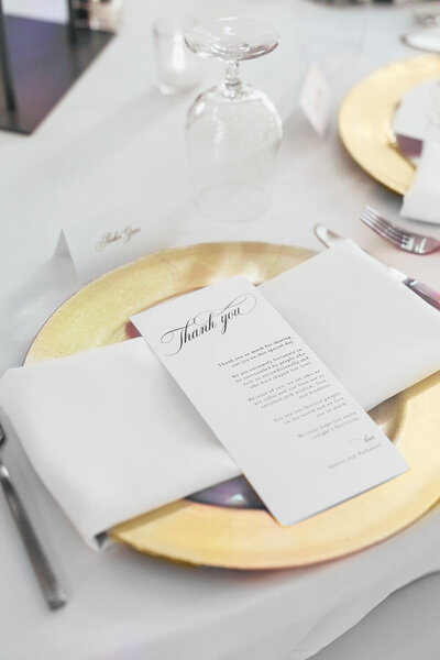 Elegant reception table setting at Ashford Acres Inn, Cynthiana KY, featuring gold chargers with white napkins and a thank you note on top, at a sophisticated black, white, and gold wedding.