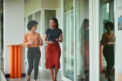 Two professional women walking through an office hallway in conversation.