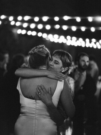 a Bride hugging a woman with her eyes closed in an intimate moment at a wedding reception with fairy lights above and behind them. This is a black and white film photo