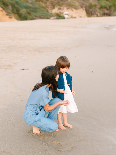candid photo of an older sister and her toddler baby sister looking at seashells at the beach