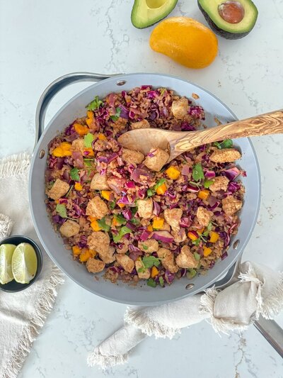 A pan of chopped chicken, with red cabbage, mango, cilantro and cauliflower rice on a marble counter.