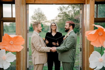 Two men stand facing each other holding hands in front of a wedding officiant. 