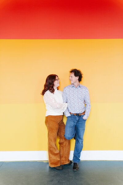 A joyful couple laughing and holding hands during a fun and colorful engagement photoshoot, surrounded by vibrant backdrops and natural light.