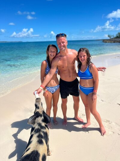 A dad and his two daughters laughing and smiling while feeding a spotted pig while on a Bahamian tour