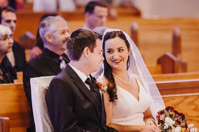 A wedding photo of Orlando Wedding Photographer Joanna from Four Loves Photo and Film, and her husband Cole. They are seated in a church pew and smiling gently at each other during the ceremony.