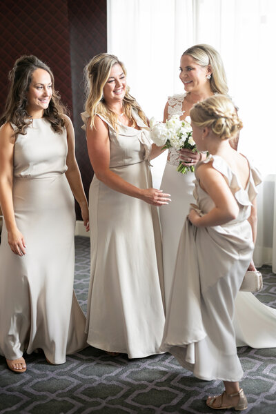 Four women in cream-colored dresses stand together, smiling and chatting. One holds a bouquet, suggesting a wedding setting. They are in a well-lit room with a patterned carpet and a window in the background.