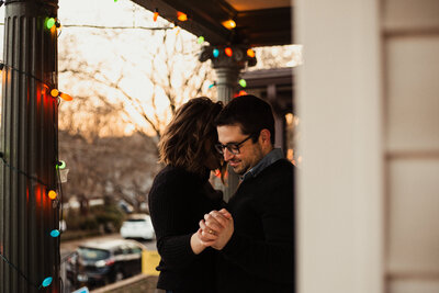 An engaged man and woman dance in autumn twilight on their front porch under brightly colored christmas tree lights.