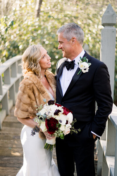 blonde woman in fur coat and white dress holding flowers looking at man in black tuxedo on a bridge 