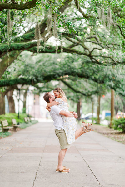 A happy man lifts his fiancee for a kiss under an oak canopy on a sidewalk