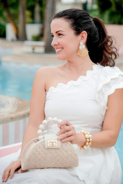 Woman wearing a one-shoulder white dress styled with statement tassel earrings, a pearl-handle bag, and stacked Gaby & Grace bracelets beside a resort pool.

