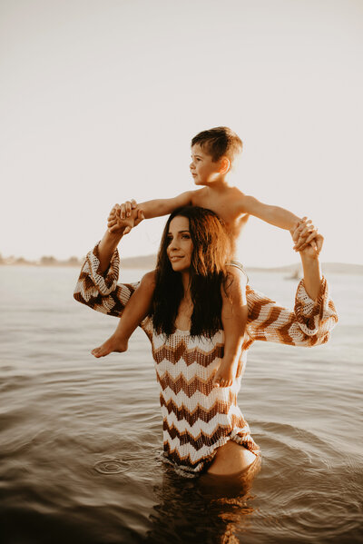 A woman stands in shallow water wearing a patterned dress, holding a young child on her shoulders. The child is shirtless, and they are both looking into the distance.ess nursing newborn near the beach