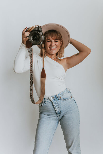 Laura, photographe de mariage en Vendée, avec un chapeau et un appareil photo, grimace joyeusement devant un fond neutre.