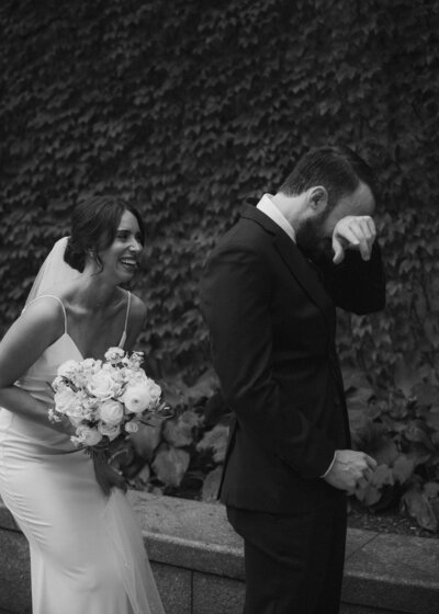 Romantic black and white photo or groom wiping his eyes while elegant bride with veil and bouquet smiles behind him for first look.
