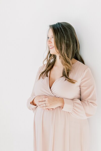 A pregnant woman in a pink dress leaning against a white wall, captured by a Charlotte maternity photographer.