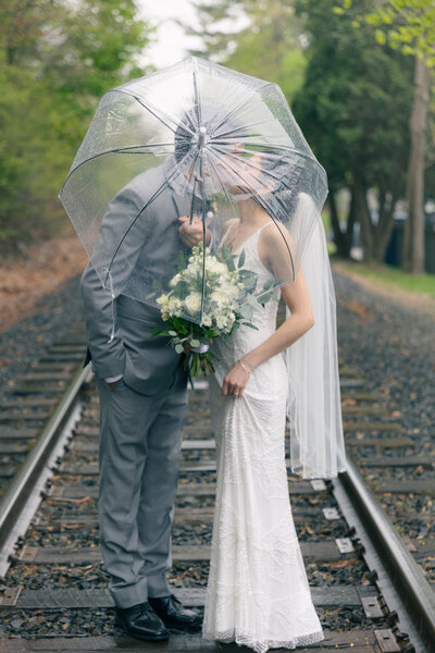 bride and groom kissing underneath a clear umbrella in the rain on abandoned traintracks