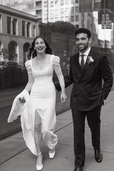 A bride and a groom walking quickly and smiling at the camera. The bride is holding her wedding dress so she can walk quickly. The groom has his hand in his pocket.