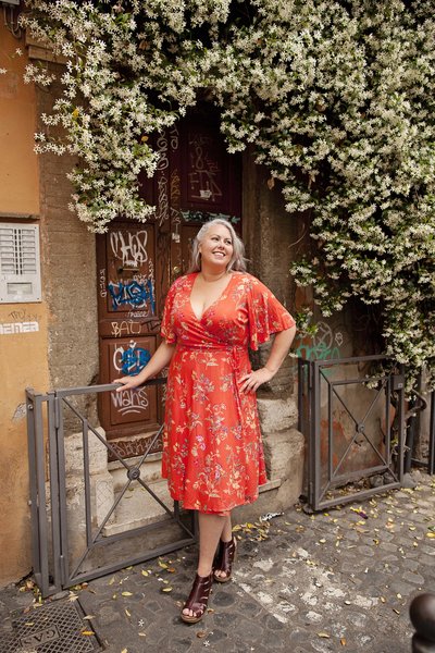 A girl in an orange and yellow floral dress standing in front of a door covered in jasmine. Taken by Rome Portrait Photographer, Tricia Anne Photography