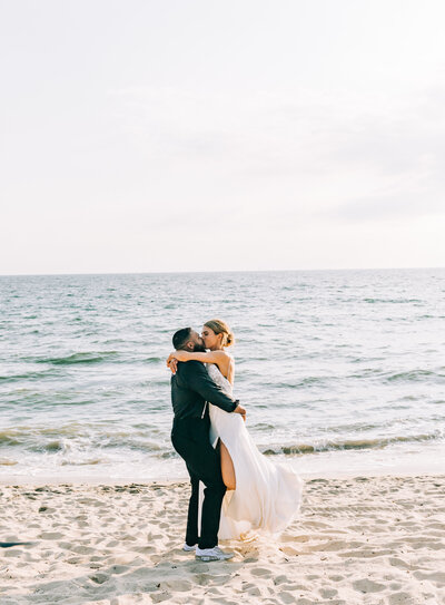 A bride and groom share an embrace on the beach, with the groom lifting the bride as they kiss. The bride wears a flowing white gown with a high slit, and the groom is dressed in dark attire. The ocean stretches out behind them under soft sunlight.