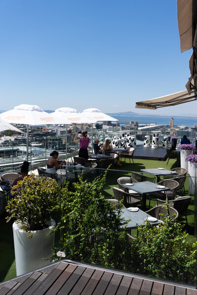 People seated at tables with umbrellas at an outdoor restaurant or cafe, suggesting a relaxed dining atmosphere in Cape Town.