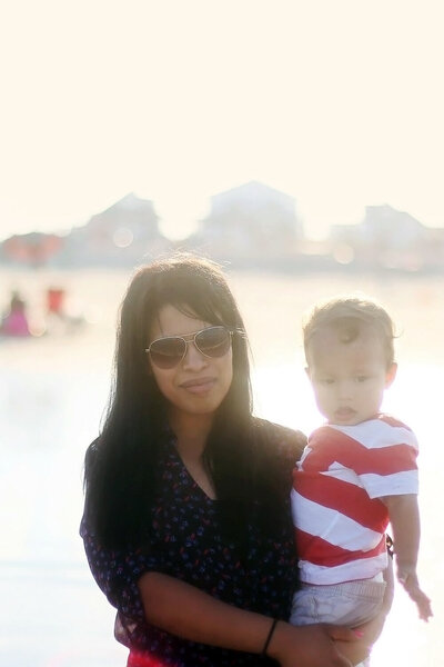 Image of mother holding young boy during sunset mother had long black hair and side swept bangs while child has red and white striped shirt.  mom is wearing sunglasses in a backlit sunset photo