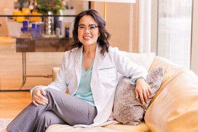 woman doctor plastic surgeon wearing white coat gray pants and light blue silk top sitting sideway on a sofa legs crossed arm resting on a pillow Laure Photography Atlanta GA