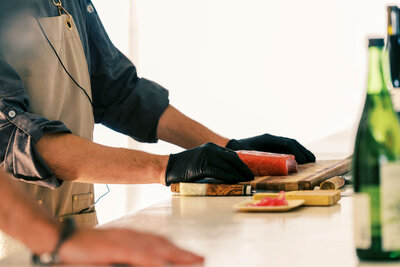 Close-up of a chef slicing sushi-grade fish on a wooden cutting board, wearing black gloves, with sake bottles in the foreground—captured by Joseph Mayden for culinary brand marketing.