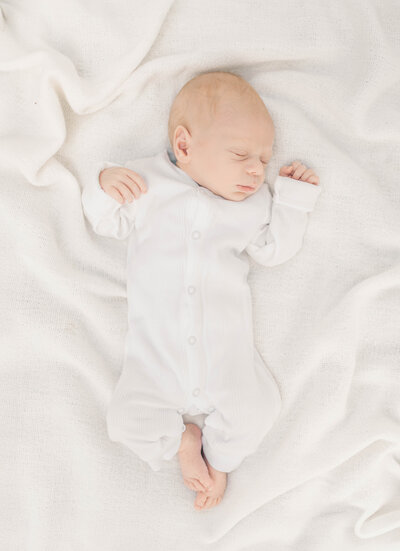 Newborn baby boy sleeping peacefully in a white sleeper on a soft blanket during a lifestyle session in Kansas City.