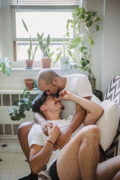 Two masculine presenting people cuddle on a couch, one lying in their partner's lap, and the other reaching down to kiss their forehead. The wall visible behind them is covered in small plants.