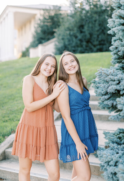 Twin senior girls lean against each other smiling at the camera while in downtown. They are wearing matching , different colored dresses.