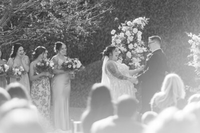a black and white photo of a bride and groom in the middle of their garden wedding. The bride has her head thrown back laughing while holding hands with the groom. 