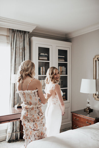 Bridesmaid in flower dress fastening buttons on back of wedding dress. Hotel room furniture, bed and night lamp.