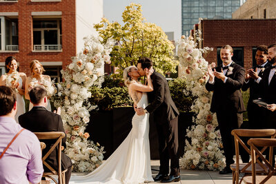 Bride in mermaid-style gown kisses groom in front of extravagant floral arch after saying "I do" in rooftop Chicago wedding.
