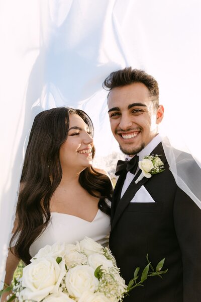 Bride and groom smiling together during their wedding portraits, bride holding white florals and groom wearing a black tux, photographed by Colorado wedding photographer at Spruce Mountain Ranch