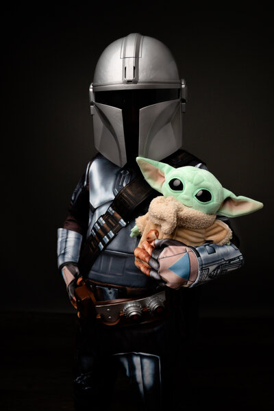 Boy dressed in Mandalorian Costume holding Grogu stuffed toy in front of black backdrop.