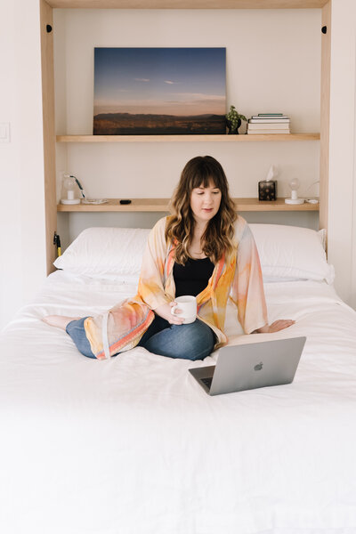 A woman sits on a bed with her laptop and a mug