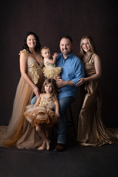 Elegant family portrait captured in a Dallas photography studio, featuring a mom and two daughters in gold and tan gowns, dad in a blue shirt with jeans, and a teenage daughter in a silky gold dress, all posed beautifully against a dark textured backdrop.